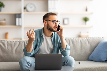 Stressed young freelancer using laptop and making call on smartphone at home office