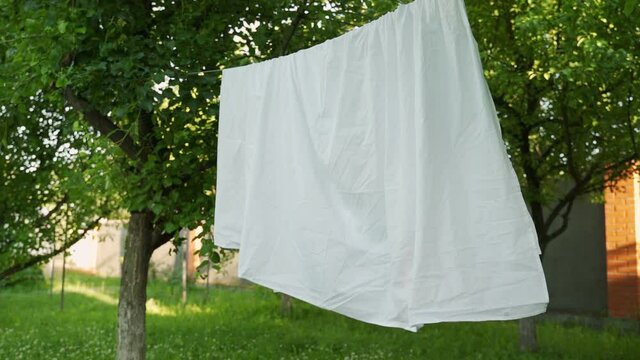 Clean white sheet is hanging on a clothesline in the backyard. Laundry is dried on a rope in the yard in the sun.