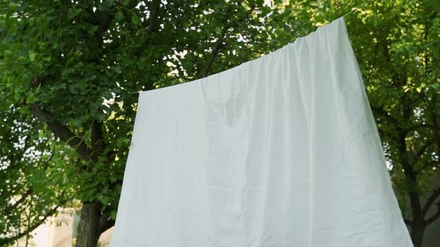 Clean white sheet is hanging on a clothesline in the backyard. Laundry is dried on a rope in the yard in the sun.
