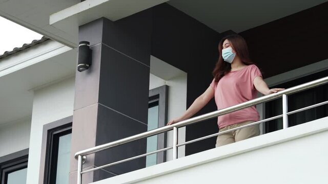 woman wearing a face mask and quarantine in balcony of her home with falling rain, coronavirus (covid-19) pandemic concept