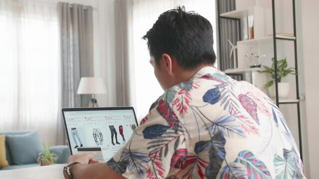 Asian Man Looks At Goods In Online Store Clothing. Buy Fashion Clothes Directly On Laptop Computer

