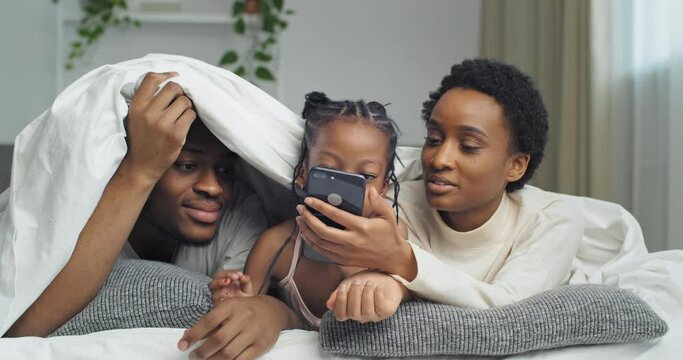 Afro american family black ethnic african parents lie on sofa bed covered with white blanket look at screen of mobile phone use wirelles smartphone to take photo watching video shopping online in net 