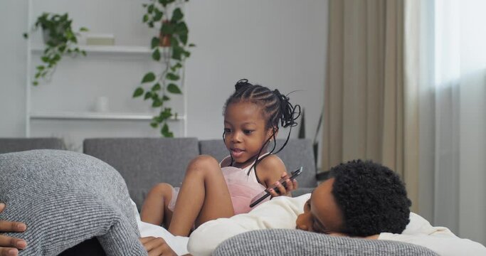 Afro American Family Black Ethnic African Parents Lying On Couch Resting Man Father Hiding Under Pillow Little Daughter Girl Child Looking At A Mobile Phone Playing Game On Smartphone Using Cellphone