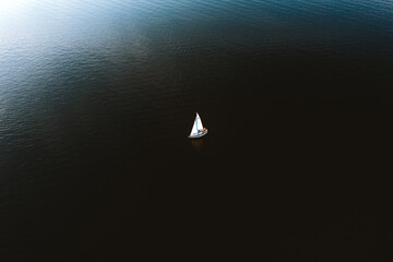 Aerial view of tiny yacht with white sails floating in calm ocean with no wind.