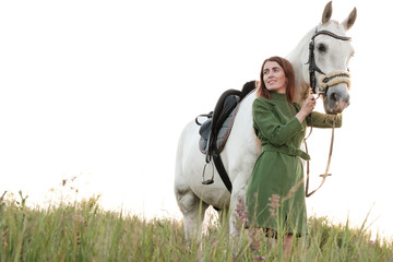 Young attractive woman in a dress is walking with her horse, holding her by the bridle, in a meadow, in the evening at sunset.