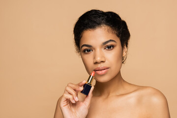 african american woman with bare shoulders holding lipstick isolated on beige