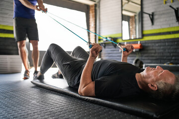 Personal trainer holding resistance bands. Elderly man does bicep curl lying on mat