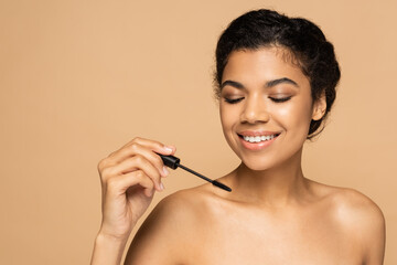 smiling african american woman with bare shoulders holding mascara brush isolated on beige
