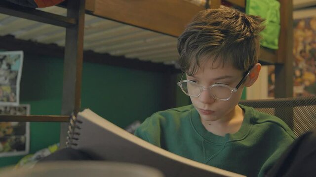 An attractive young boy drawing in a sketchbook in his bedroom pauses to look out the window.