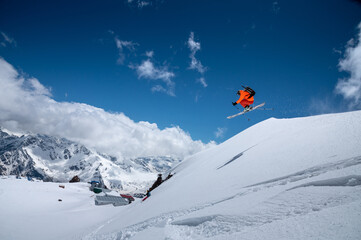 Naklejka na ściany i meble Professional athlete young male skier in an orange ski suit flies over the mountains after jumping from snow-covered ledges. Freeride sports community against the backdrop of snow-capped peaks