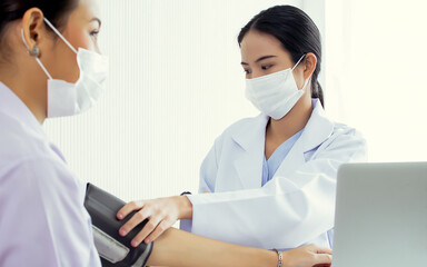 Portrait smart Asian female young doctor wearing face mask to protect virus, white gown coat, measuring blood pressure and giving advice to patient at hospital. Medical Treatment Concept.