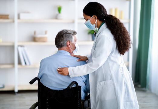 Young Female Doctor In Face Mask Listening To Old Man's Breathing, Using Stethoscope At Home