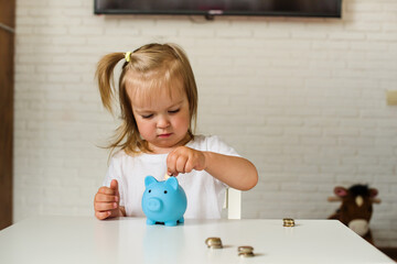 Little Caucasian girl putting coins in to the pig-shaped money box, blue piggy bank. The child...