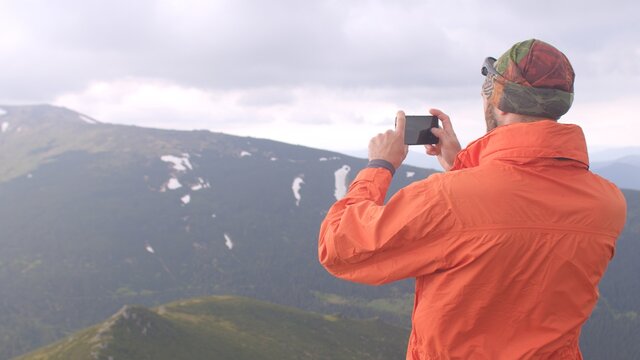 A guy in orange clothes is taking pictures of a mountain valley. He uses the phone in his hands, directs and takes a picture. Preservation of impressions in the image.