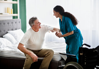 Caregiver helping elderly male to get into wheelchair at home. Rehabilitation and medical care for...