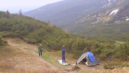 A tourist does a body workout in a camp in the mountains. A man comes up with water, communication of friends. Against the background of the mountain. Distance from everyday life.