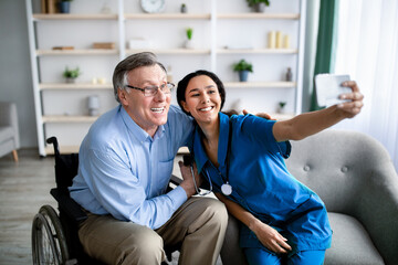 Obraz premium Cheerful impaired senior man in wheelchair taking selfie with young female doctor at retirement home