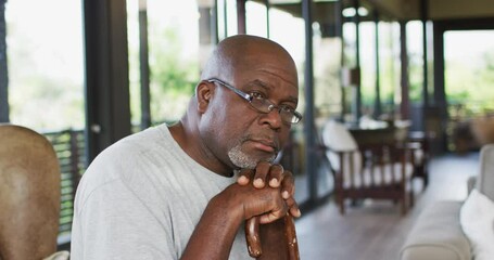 Portrait of thoughtful african american senior man sitting in living room leaning on walking stick - Powered by Adobe