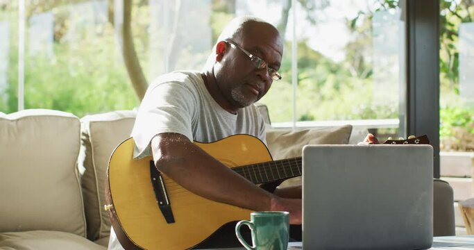 Happy african american senior man in living room playing acoustic guitar and using laptop