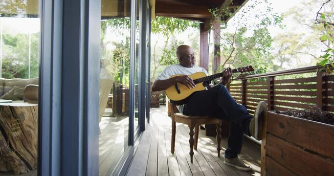 Thoughtful african american senior man sitting on balcony playing acoustic guitar
