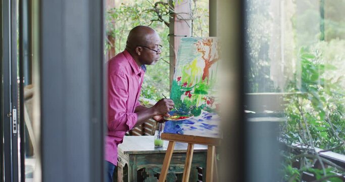 Happy african american senior man standing on balcony painting picture of the view