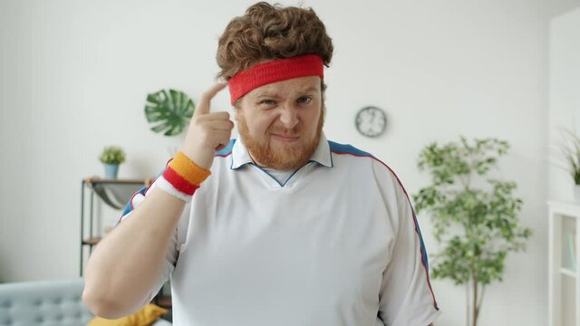Slow motion of silly obese guy playing with sports headband then showing fool hand gesture standing indoors at home and looking at camera. Emotions and sportsman concept.
