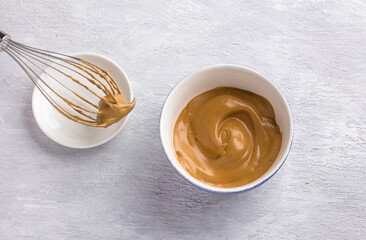 Ceramic bowl and whisk with fluffy creamy whipped coffee foam on a light gray textured background. For dalgona coffee, trendy food and drink concept