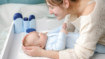 Obraz premium Smiling mother stroking and smiling at her little baby boy lying on dressing table at bedroom. Concept of hygiene, baby care and healthcare