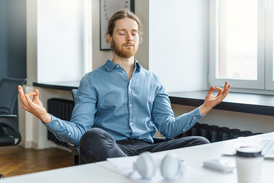 Taking break. Calm and peaceful young businessman with closed eyes meditating and relaxing during hard working day, keeping hands in mudra gesture while sitting in lotus pose at work in office