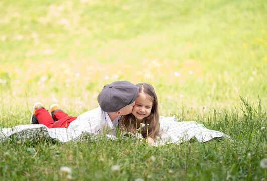 Children lie on a blanket in a green field. The boy kisses a little girl on the cheek.