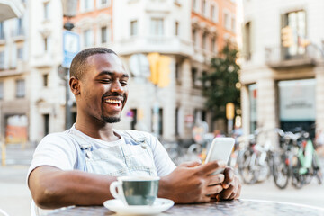 Attractive afro american man sitting at a bar terrace. He is using his phone and smiles.