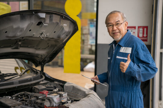 Professional senior car mechanic giving thumb up near automobile working in the garage workshop. Elderly vehicle mechanic holding tablet maintenance the engine in an auto repair service center.