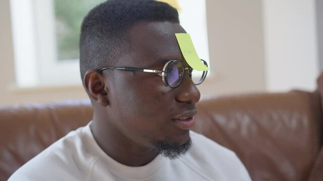 Close-up of African American millennial man guessing Stone word playing with friends indoors. Portrait of handsome cheerful guy in eyeglasses talking in slow motion