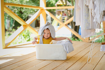 cute caucasian toddler girl in a basket with clean linen on the veranda, hangs up the washed, and...