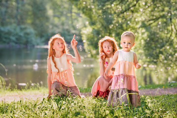 Three red-haired sisters in long linen dresses blow bubbles in the park on sunny summer day.