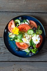 Appetizing salad with feta cheese, fresh vegetables (tomatoes, cucumbers, onions), crispy lettuce, microgreens and violet flowers.  Greek salad on a wooden background. Food with flowers