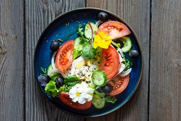 Appetizing salad with feta cheese, fresh vegetables (tomatoes, cucumbers, onions), crispy lettuce, microgreens and violet flowers.  Greek salad on a wooden background. Food with flowers