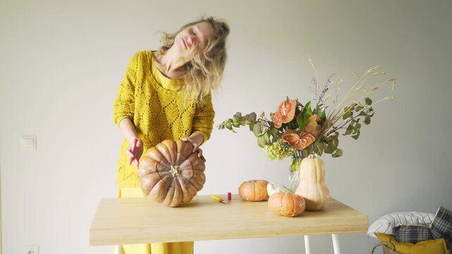 Pretty blondie woman in yellow sweater holding large orange pumpkin in hands while drawing with red marker to make traditional halloween decor in light room at grey wall solid background