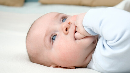 Portrait of adorable baby boy with blue eyes sucking his hand while lying on bed