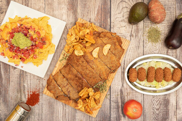 Top view image of food with the main dish of cachopo with potatoes, cod croquettes and nachos with guacamole.