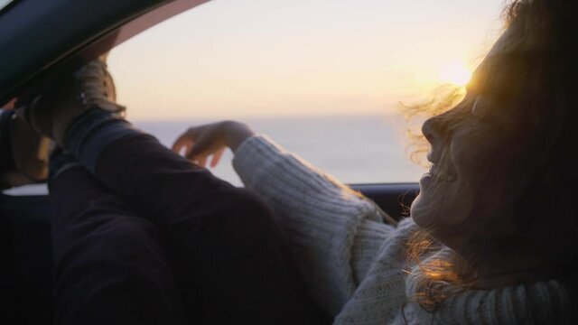 Young woman enjoys soft wind with legs stuck out of modern automobile open window on tranquil ocean beach with sun path at bright sunset light close view