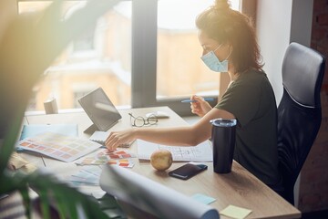 Side view of interior designer or architect wearing protective mask selecting color from samples while working on new poject, sitting at the table in her office
