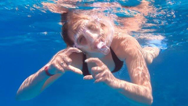 Young woman in a mask with a snorkel swimming underwater, smiles and shows a heart sign with her hands