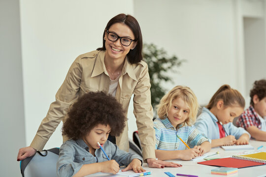 Attractive young female teacher in glasses smiling at camera while helping little students. Kids sitting at the table, studying in elementary school