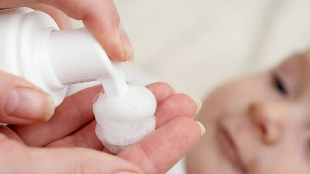 Closeup of applying medication lotion on red baby cheek suffering from dermatitis and acne. Concept of newborn baby hygiene, health and skin care