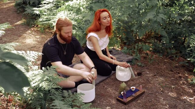 Red-haired couple playing on white quartz crystal singing bowls in the forest. Healing medicine and meditation Spiritual music Buddha