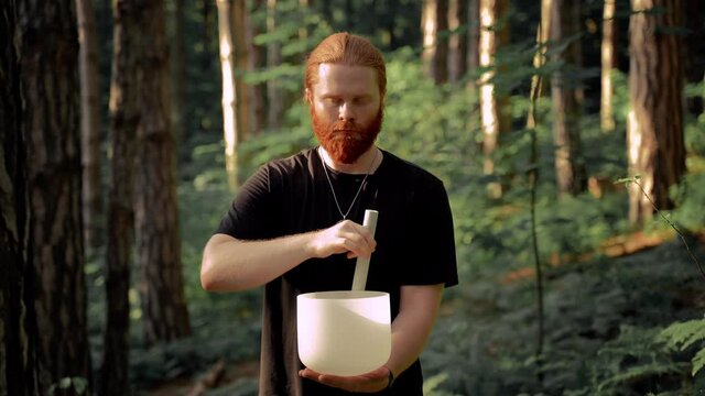 Red-haired man playing on white quartz crystal singing bowls in the forest. Healing medicine and meditation Spiritual music Buddha