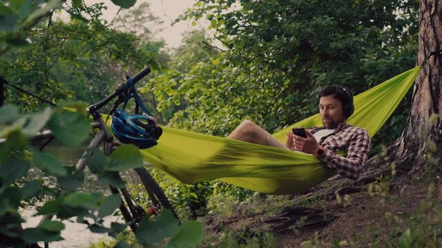 Man on bicycle trip at camping by lake is relaxing in green hammock while listening to music. Active recreation theme in nature. Hipster cyclist with headphones having fun in hammock by river.