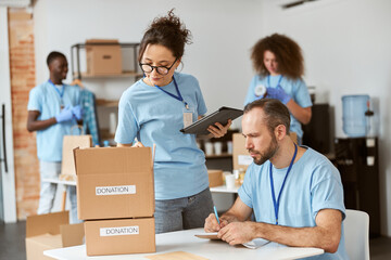 Man and woman working together on donation project indoors. Team of volunteers in blue uniform making notes, calculating, sorting and packing items in cardboard boxes