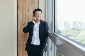 Happy and smiling Asian businessman talking on the phone in a modern office center, sharing good news with companions